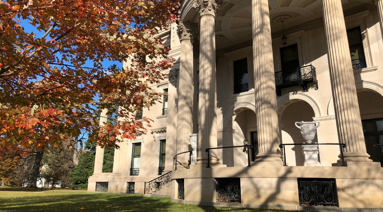 Vanderbilt Mansion in Hyde Park, with its stone columns, is photographed on a sunny autumn day with a tree with brown leaves in the foreground to the left.